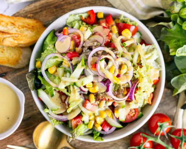 Ein bunter gemischter Salat mit knackigem Salat, Tomaten, Gurken, Mais und roten Zwiebeln, serviert mit Dressing und geröstetem Brot.