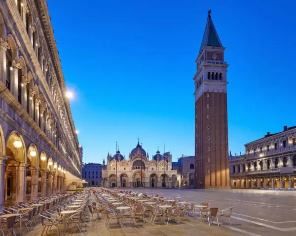 Piazza San Marco a Venezia, con la Basilica e il Campanile, testimoni della ricca storia della città. Tavolini all'aperto creano un'atmosfera serale.