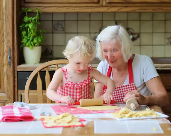 Babcia z wnuczką pieką ciasteczka. Dziewczynka wałkuje ciasto, a babcia z uśmiechem patrzy na jej pracę.