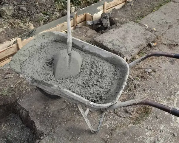 Taczka z betonem i łopatą czeka na użycie przy budowie fundamentów. W tle widać szalunki i przygotowane zbrojenie.