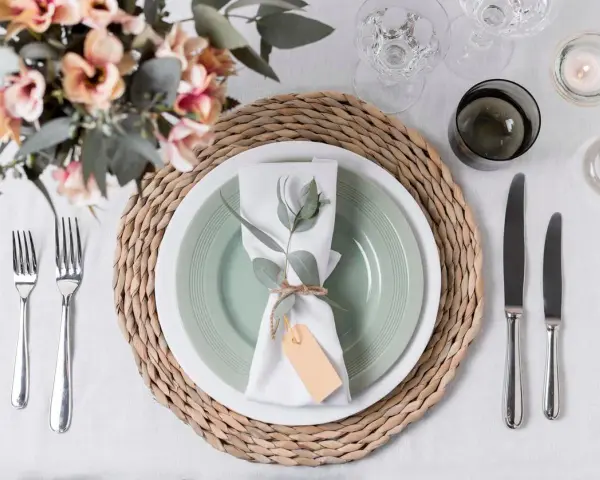 Une table dressée avec une assiette verte, une serviette blanche décorée d'une branche d'eucalyptus et d'une étiquette, le tout sur un rond de serviette fait maison.