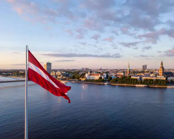 Flaga Łotwy powiewa nad rzeką w Rydze, ukazując panoramę miasta o zachodzie słońca.