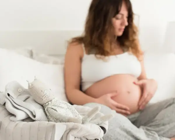 Czekając na pierwsze kopnięcia, przyszła mama głaszcze brzuszek. Obok leżą ubranka i przytulanka dla maluszka.