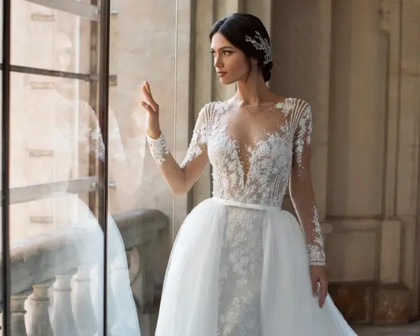 Panna młoda w sukni ślubnej z koronką i trenem, z ozdobną fryzurą w stylu bollywood, patrzy przez okno.