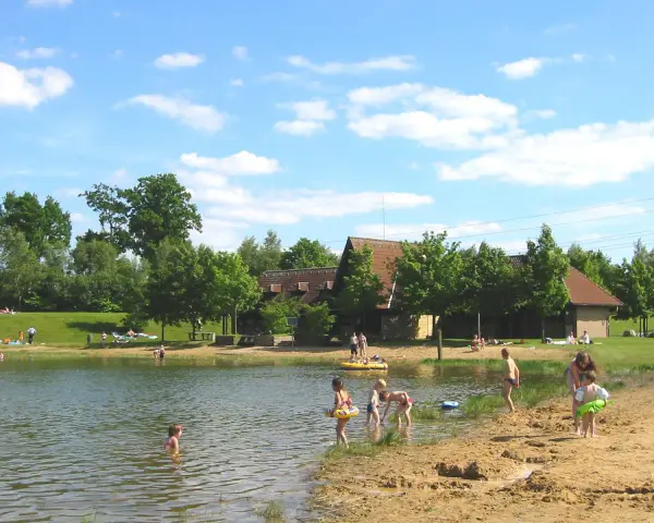 Sonniger Tag am badesee fredenbeck. Kinder planschen im Wasser und am Sandstrand, während im Hintergrund Gebäude und Bäume zu sehen sind.