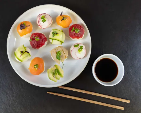 Na talerzu ułożone są kolorowe kulki sushi z łososiem, wołowiną, awokado i rzodkiewką. Obok miseczka sosu sojowego i pałeczki.