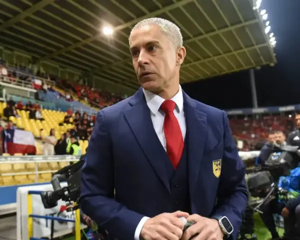 Sylvinho, trener w granatowym garniturze i czerwonym krawacie, stoi na tle trybun stadionu z flagą Chile.