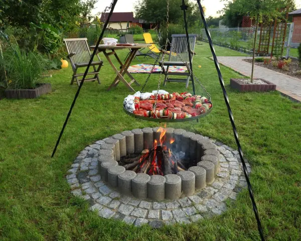 Ognisko w kamiennym kręgu, nad którym wisi okrągła taca z mięsem i warzywami na szaszłykach, gotowe do grillowania w ogrodzie.