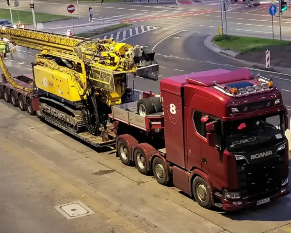 Czerwona ciężarówka Scania z przyczepą transportuje żółtą maszynę wiertniczą. Pilot pojazdu nienormatywnego czeka na przejazd.