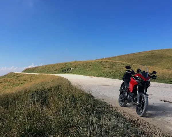 Ducati rossa ferma su strada sterrata, con un passo branchetto sullo sfondo e cielo azzurro.