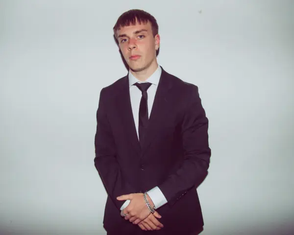 Młody mężczyzna w czarnym garniturze i białej koszuli, z krawatem. Kinny Zimner prezentuje się elegancko.
