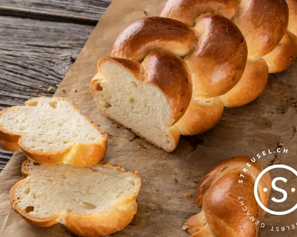 Ein goldbraunes Zopf Brot, frisch gebacken, liegt auf Backpapier. Zwei Scheiben sind abgetrennt und zeigen die weiche Krume.