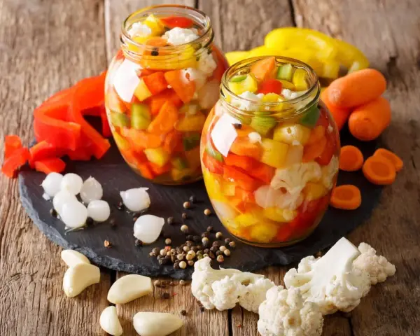 Due barattoli di giardiniera fatta in casa, pieni di verdure colorate come peperoni, carote e cavolfiore, su un tagliere di ardesia con ingredienti freschi intorno.