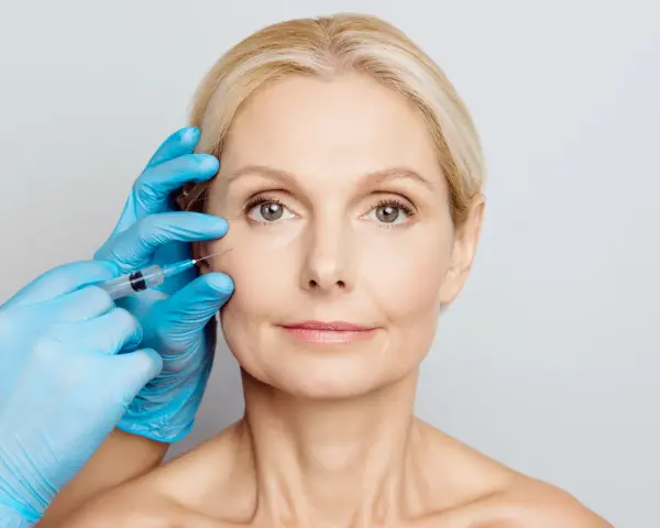 Zabieg z użyciem botoksu, czy botox jest bezpieczny? Dłoń w niebieskiej rękawiczce wykonuje zastrzyk w okolicę oka kobiety.