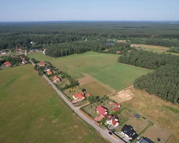 Widok z lotu ptaka na wieś otoczoną lasem. Widać domy z czerwonymi dachami, pola uprawne i jezioro. Każdy dom zajmuje swój kawałek ziemi, a całość tworzy malowniczy krajobraz.