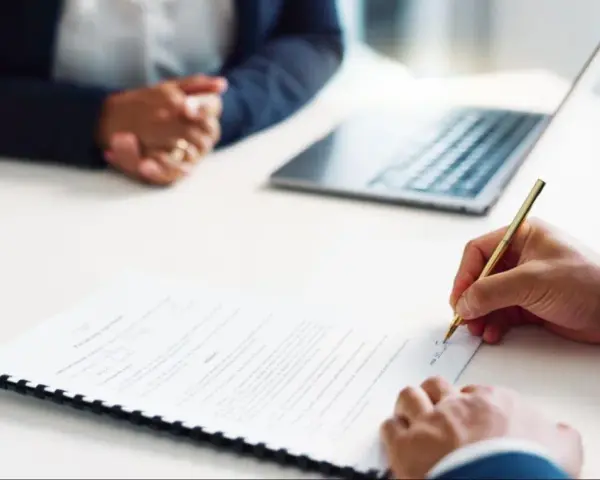 Persona firma un contrato de alquiler de habitación. A su lado, otra persona espera, con un portátil y una tablet sobre la mesa.