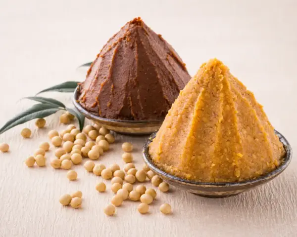 Dwie góry pasty miso, ciemna i jasna, obok ziaren soi i liści. Poznaj składniki do przygotowania tej aromatycznej pasty miso.