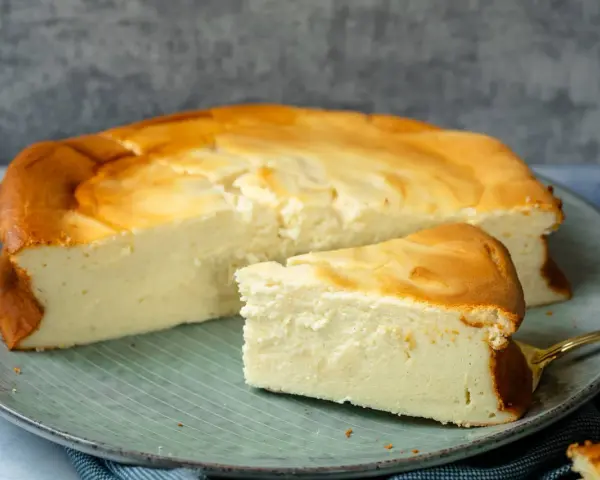 Ein saftiger Quarkkuchen ohne Boden, goldbraun gebacken, liegt auf einem Teller. Ein Stück ist bereits angeschnitten und zeigt die cremige Konsistenz.