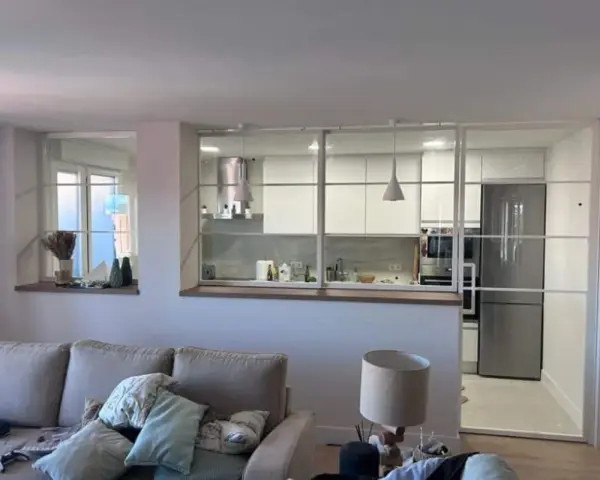 Un salón moderno con sofá y lámpara, separado de una cocina blanca y luminosa por una cristalera.