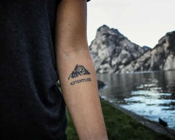 Tatuaż góry na przedramieniu z napisem "ADVENTURE". Symbolizuje miłość do podróży i odkrywania nowych miejsc.