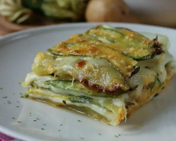 Una fetta di parmigiana di zucchine, stratificata con formaggio filante e zucchine grigliate, servita su un piatto bianco.