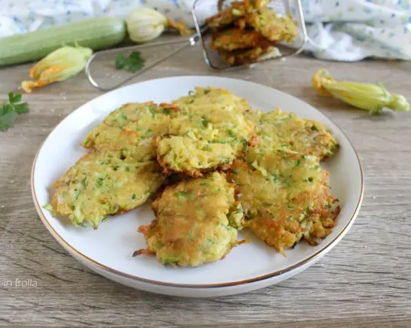 Buñuelos de calabacín dorados y crujientes en un plato blanco, con calabacines y flores en el fondo.
