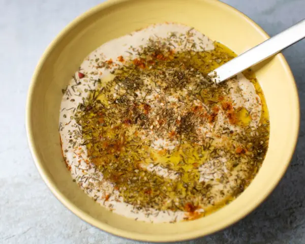 Cremiger Hummus Aufstrich in gelber Schüssel, beträufelt mit Olivenöl und bestreut mit Kräutern und Paprika.