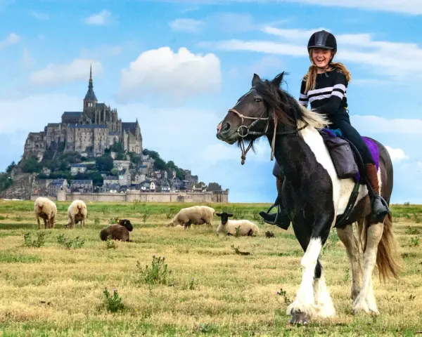 Une jeune fille rit à cheval devant le Mont-Saint-Michel, avec des moutons dans un pré.