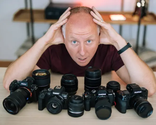 Mężczyzna z rękami na głowie otoczony aparatami różnych marek: Sony, Canon, Fujifilm.