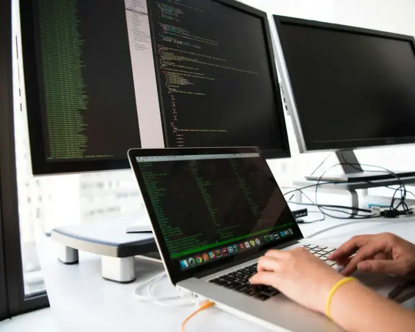 Hands typing on a laptop, surrounded by monitors displaying code. This setup is ideal for developing network infrastructure software.