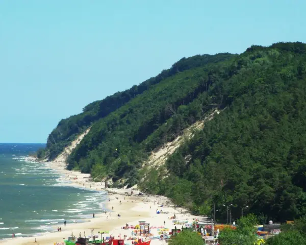 Słoneczna plaża u podnóża zalesionej Kawczej Góry. Woda faluje, a na piasku odpoczywają ludzie.