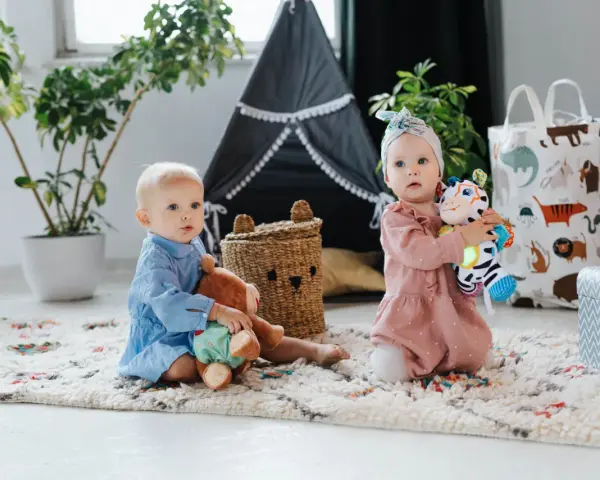 Dwoje dzieci bawi się na dywanie. Jedno trzyma misia, drugie kolorową zebrę. Idealne zabawki, co kładziemy dziecku na roczek.