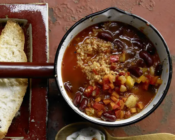 Herzhaftes Chili sin Carne mit Kidneybohnen, Bulgur und Gemüse in einem Topf, daneben geröstetes Brot.