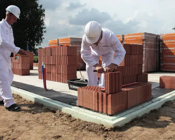 Dwóch budowlańców w białych kombinezonach układa cegły na fundamencie.