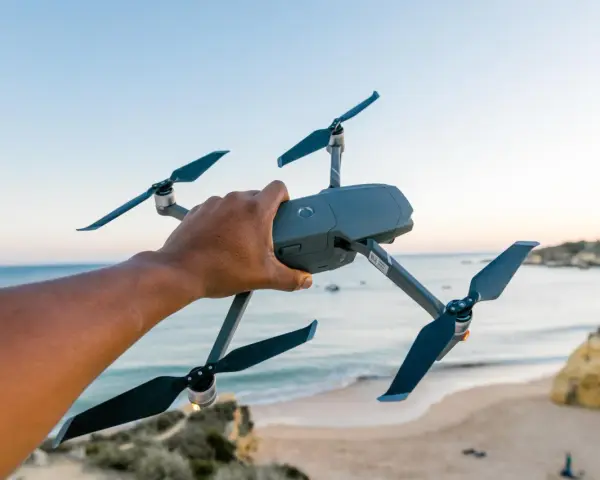 A hand holds a drone over a European beach, ready for flight.
