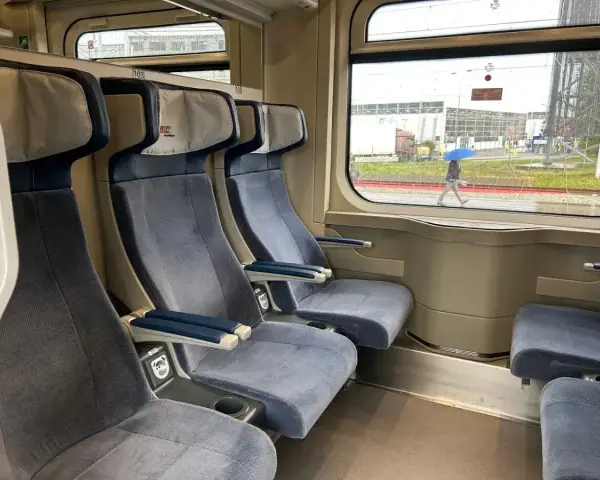 Wygodne siedzenia w wagonie z przedziałami Intercity 2 klasa. Za oknem deszczowa pogoda i przechodzień z niebieskim parasolem.