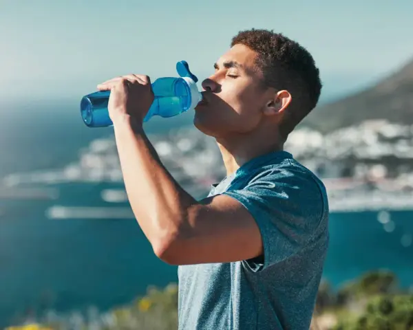Un jeune homme boit dans une gourde bleue, se réhydratant après l'effort. L'eau, essentielle, contient des électrolytes pour le bon fonctionnement du corps.