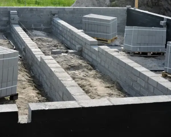 Budowa fundamentów z bloczków betonowych. Widać ułożone ściany fundamentowe, piasek w wykopie i palety z bloczkami.
