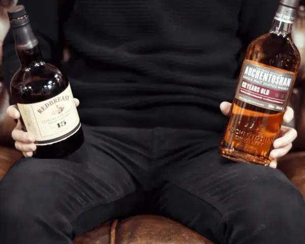 Mann hält Flaschen von Redbreast (irischer Whiskey) und Auchentoshan (schottischer Whiskey), um den Unterschied zu zeigen.