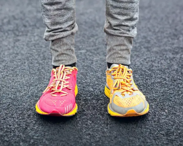 Gambe con pantaloni grigi e scarpe da corsa, una rosa acceso e l'altra arancione, con lacci gialli. La suola scarpa è pronta per correre.