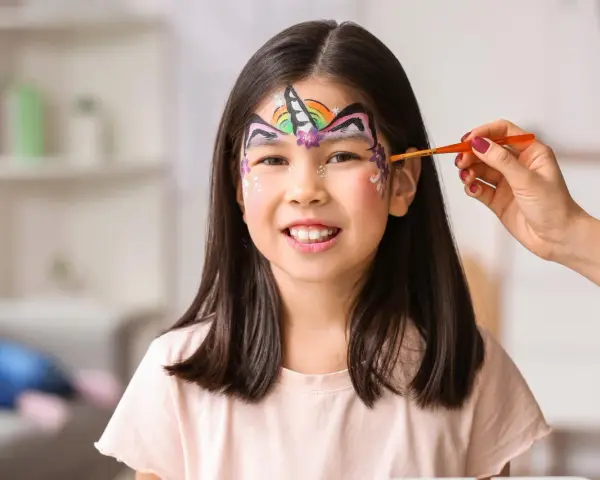 Ein Kind wird mit einem bunten Regenbogen-Einhorn-Gesicht bemalt. Ein Pinsel wird sanft über die Stirn geführt.