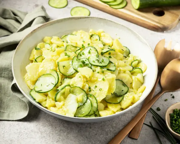 Schüssel mit Kartoffelsalat mit Gurken, garniert mit Schnittlauch. Daneben liegen Servierbesteck und eine kleine Schale mit gehacktem Schnittlauch.