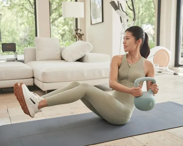 Kobieta wykonuje ćwiczenia na brzuch z kettlebell, siedząc na macie w domowej siłowni.