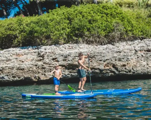 Zwei Jungen auf SUP-Boards, die geradeaus fahren, genießen das klare Wasser vor einer felsigen Küste mit üppiger Vegetation.