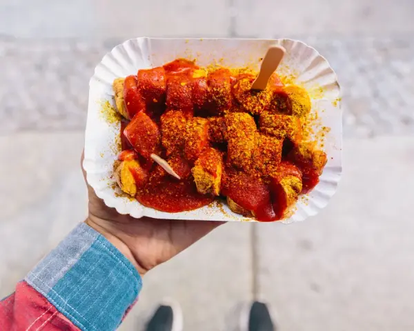 Eine Hand hält eine Portion Currywurst mit original Currywurst Sauce, bestreut mit Currypulver. Ein Holzspieß steckt darin.