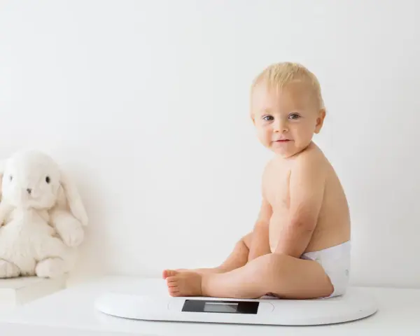 Uśmiechnięty chłopczyk w pieluszce siedzi na wadze. Chyba już czas na większy rozmiar, kiedy pampers jest za mały.