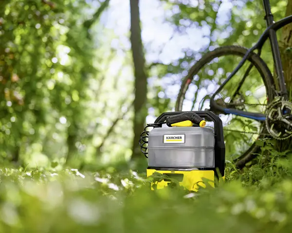 En Kärcher OC3 portabel rengörare står i gräset bredvid en cykel lutad mot ett träd. Perfekt för att snabbt skölja av utrustning efter ett äventyr.
