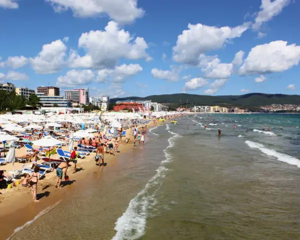 Tłum ludzi wypoczywa na piaszczystej plaży Słoneczny Brzeg, opalając się na leżakach pod białymi parasolami. W tle widać morze i zabudowania kurortu.