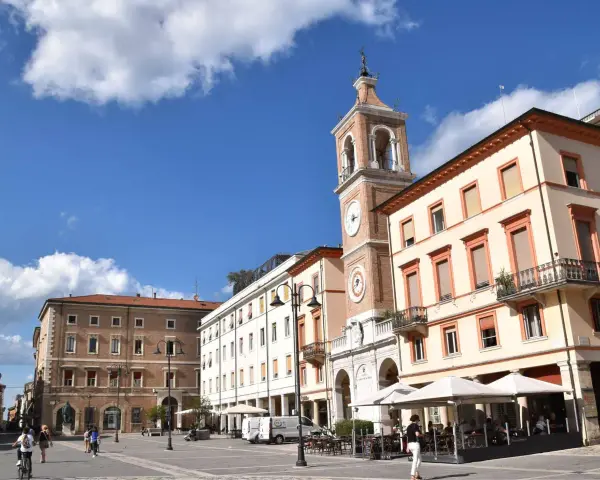 Piazza Tre Martiri a Rimini: un campanile con orologio domina la scena, tra palazzi storici e tavolini all'aperto. Ecco cosa vedere!