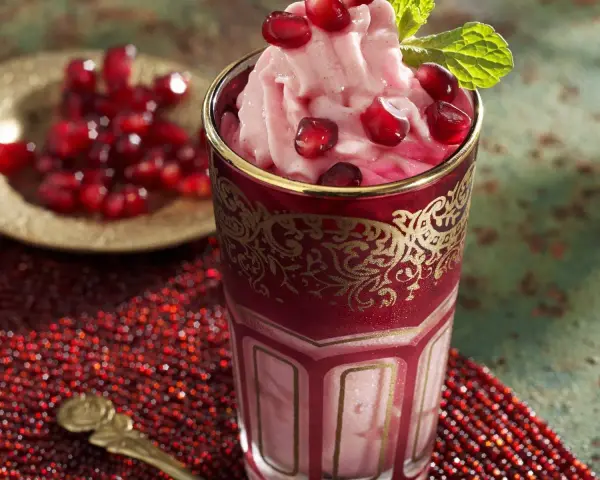 Ein Glas mit cremigem granatapfel eis, garniert mit frischen Granatapfelkernen und Minze. Daneben eine Schale mit Granatapfelkernen.